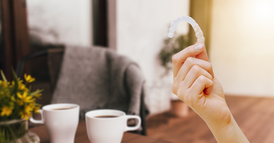 A person holding up an Invisalign aligner next to two hot beverages.